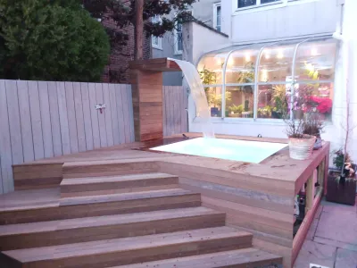 A custom wooden deck features wide steps leading up to a small spa with a modern waterfall, situated next to a glass-enclosed sunroom.