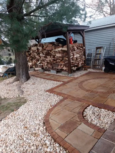 A backyard scene featuring a curved paver patio bordered by white gravel leading past a large tree and a covered, stacked firewood shed.