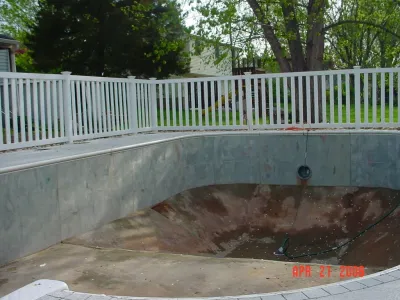 A view of an empty residential swimming pool shell undergoing construction, featuring raw concrete walls and a surrounding white vinyl safety fence.
