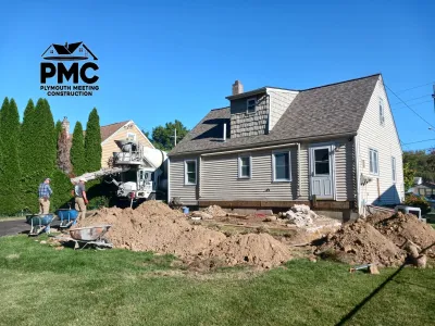 Two construction workers use a cement truck and wheelbarrows to pour concrete for a residential home foundation, surrounded by piles of excavated dirt.